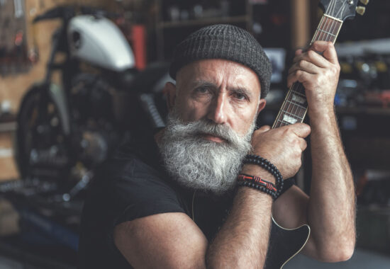 Assured mature man keeping instrument Confident bearded biker is siting in garage and holding black guitar. He glancing at camera with expectation. Portrait